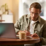 رجل يعمل بتركيز على جهاز الكمبيوتر مع دفتر ملاحظات وكوب قهوة في أجواء هادئة. تعبر الصورة عن مفهوم مواقع العمل الحر والعمل عن بعد من أي مكان، مما يتيح المرونة وزيادة الدخل من خلال منصات العمل المستقل.