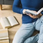 صورة لشخص يجلس على الأرض ويرتدي سترة زرقاء، يقرأ كتابًا محاطًا بعدة كتب مفتوحة. تعكس الصورة شغف القراءة والاستغراق في المعرفة.