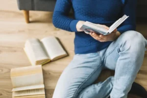 صورة لشخص يجلس على الأرض ويرتدي سترة زرقاء، يقرأ كتابًا محاطًا بعدة كتب مفتوحة. تعكس الصورة شغف القراءة والاستغراق في المعرفة.