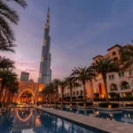 صورة ساحرة لبرج خليفة، أطول برج في العالم، أثناء غروب الشمس في مدينة دبي. يظهر البرج محاطًا بالنخيل والمباني العريقة مع انعكاس رائع على بركة المياه في المقدمة. تُبرز الصورة جمال العمارة الفاخرة وروعة المناظر في دبي.