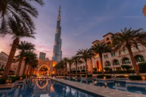صورة ساحرة لبرج خليفة، أطول برج في العالم، أثناء غروب الشمس في مدينة دبي. يظهر البرج محاطًا بالنخيل والمباني العريقة مع انعكاس رائع على بركة المياه في المقدمة. تُبرز الصورة جمال العمارة الفاخرة وروعة المناظر في دبي.